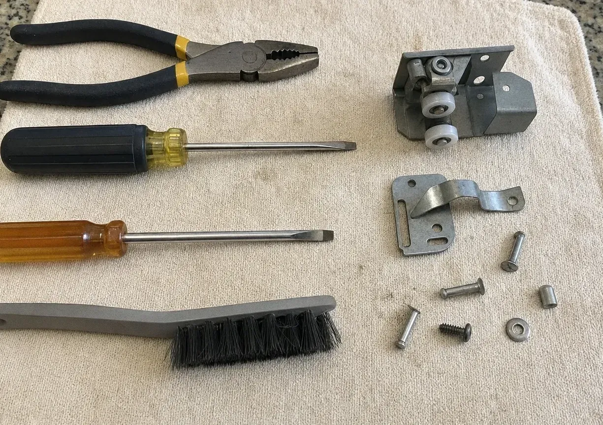 Lineman's pliers, two screwdrivers and a wire brush laid out beside a roller bracket, a steel bracket and loose bolts